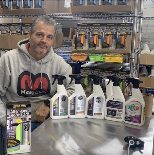 Tom Munro, Founder of Supreme Surface® and Munro-Co Brands. Tom's sitting at a stainless steel table with a bottle of Supreme Surface Granite Quartz & Marble Treatment, Daily Stone Cleaner, Scum & Mineral Deposit Remover, and NuVibrants Composite Granite Sink Treatment.